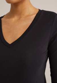 Juodas V-kaklo ilgomis rankovėmis marškinėliai, pagaminti iš lygios, elastingos medžiagos su prigludusia forma. Yra gili iškirptė ir tvarkingi siuvimo akcentai.