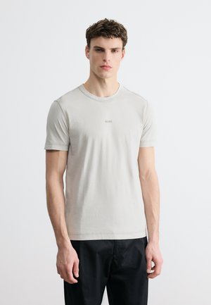 Νεαρός άνδρας με ανοιχτό γκρι T-shirt BOSS και μαύρο παντελόνι, στέκεται μπροστά σε απλό λευκό φόντο.