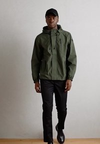 Green waterproof jacket with a high collar, adjustable hood, and zippered pockets, worn over black pants and black shoes.
