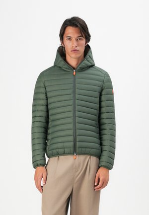 Jonge man in een groene gequilte hoodie met ritssluiting, staande tegen een plain witte achtergrond, in beige broek.