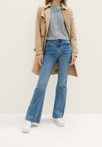 Beige trenchcoat over en stribet skjorte med knapper, parret med flagrende blå jeans og hvide sneakers. Glatte teksturer, afslappet pasform.