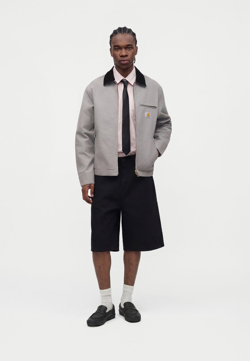 Mann trägt eine hellgraue Jacke mit Reißverschluss und schwarzem Kragen, rosa Hemd, schwarze Krawatte, schwarze knielange Shorts, weiße Socken und schwarze Schuhe, steht still.