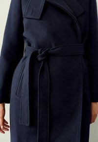 Manteau en laine bleu marine avec un large col, des manches longues et une ceinture nouée à la taille ; texture lisse avec des poches latérales et des coutures ajustées.