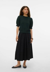 Pull léger à manches courtes de couleur vert foncé porté avec une longue jupe noire. Texture lisse, coupe décontractée et un léger éclat. Des ballerines noires à motifs complètent la tenue.