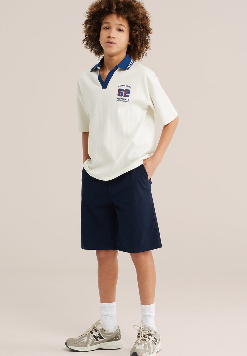 Garçon aux cheveux bouclés portant un polo blanc avec un col bleu et le numéro 62, un short bleu marine, des chaussettes blanches et des baskets beiges, debout les mains dans les poches.