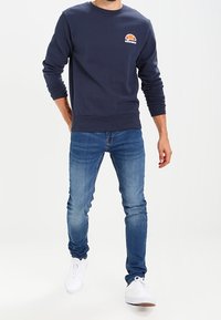 Sweatshirt bleu marine avec des poignets côtelés, col rond et logo brodé sur la poitrine gauche, associé à un jean skinny bleu clair et des baskets blanches.