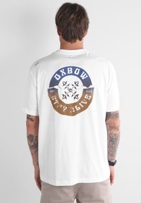 T-shirt en coton blanc avec un graphique circulaire aux accents bleus et marron, "OXBOW", des plantes, et "EST. 1985" imprimé au dos.