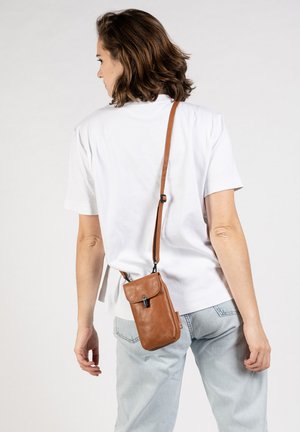 Persoon draagt een wit shirt met korte mouwen en lichtblauwe jeans, en draagt een kleine bruine leren crossbodytas met klep en sluiting.