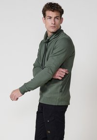 Hombre ajustando la manga de una sudadera texturizada verde oliva con cordones negros, vistiendo pantalones cargo negros, mirando hacia la izquierda contra un fondo gris liso.