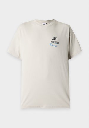 T-shirt Nike beige clair à manches courtes, col rond, petit logo "Nike Arts Club" et motif bleu sur la poitrine gauche.