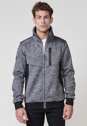 Chaqueta gris y negra con cremallera, cuello alto, tejido texturizado, detalles en negro y un bolsillo con cremallera en el pecho.