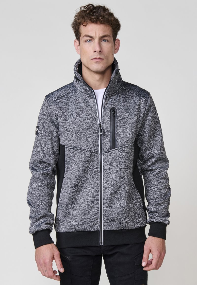 Chaqueta gris y negra con cremallera, cuello alto, tejido texturizado, detalles en negro y un bolsillo con cremallera en el pecho.