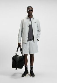 Grijze button-up shirt over een grijze T-shirt, gecombineerd met lichtgrijze shorts. Zwarte sportschoenen en een zwarte sporttas. Donkere zonnebril.