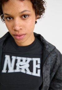 Črn Nike majica s poudarjenim belim logotipom. Model nosi temno vzorčasto jakno, kar daje mehak in sproščen videz.