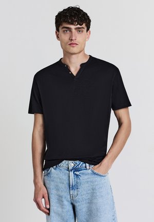 Schwarzes T-Shirt mit kurzen Ärmeln, das einen Henley-Kragen mit zwei Knöpfen aufweist und aus einem strukturierten Stoff gefertigt ist. Es wird mit hellen Denim-Shorts kombiniert.