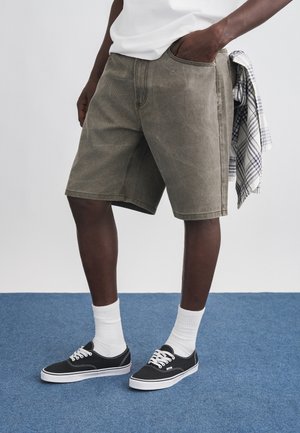 Mees, kandes pleekinud oliivivärvi lühikesi pükse, valgeid keskmise pikkusega sokke, musti lõuendjalatseid ja valget särki, hoides vöökoha juures ruudulist särki.