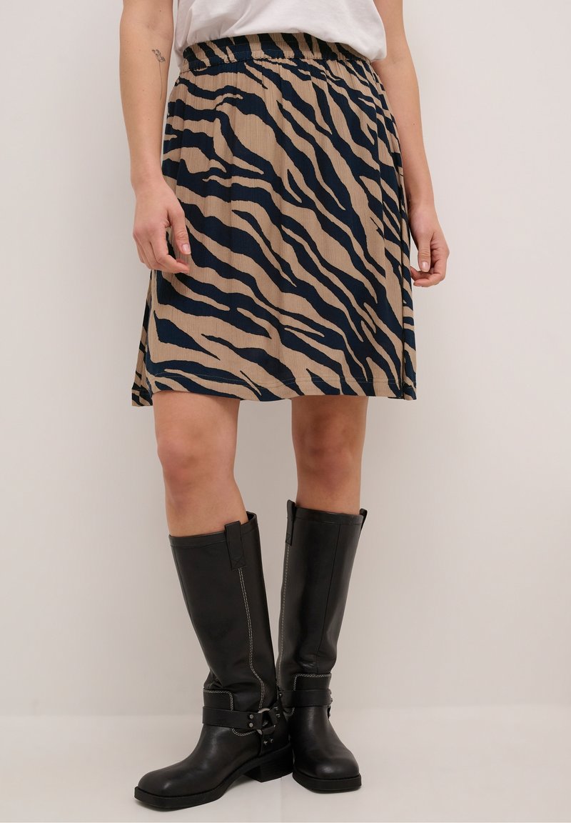 Femme portant une jupe beige et noire à motif zèbre avec des bottes en cuir noires montantes jusqu'aux genoux, debout contre un mur clair uni.