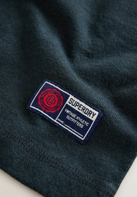 Tume mereka sviitsi, millel on sinise ja punase värvikombinatsiooniga silt, kus on õmmeldud tekst "SUPERDRY" ja "VINTAGE ATHLETIC OUTFITTERS" hemmi juurde.