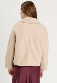 Veste en mélange de laine beige avec un large col, des épaules tombantes et une coupe boxy, assortie d'une jupe fluide bordeaux.