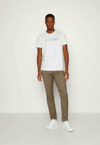 Λευκό βαμβακερό μπλουζάκι με το λογότυπο "HILFIGER", συνδυασμένο με καφέ στενά παντελόνια και λευκά αθλητικά παπούτσια. Η απλή φόντο αναδεικνύει το σύνολο.