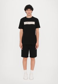 Svart bomull T-skjorte med en kremfarget logopanel, iført med svarte shorts. Antrekket er kombinert med hvite joggesko og er stylet casual.