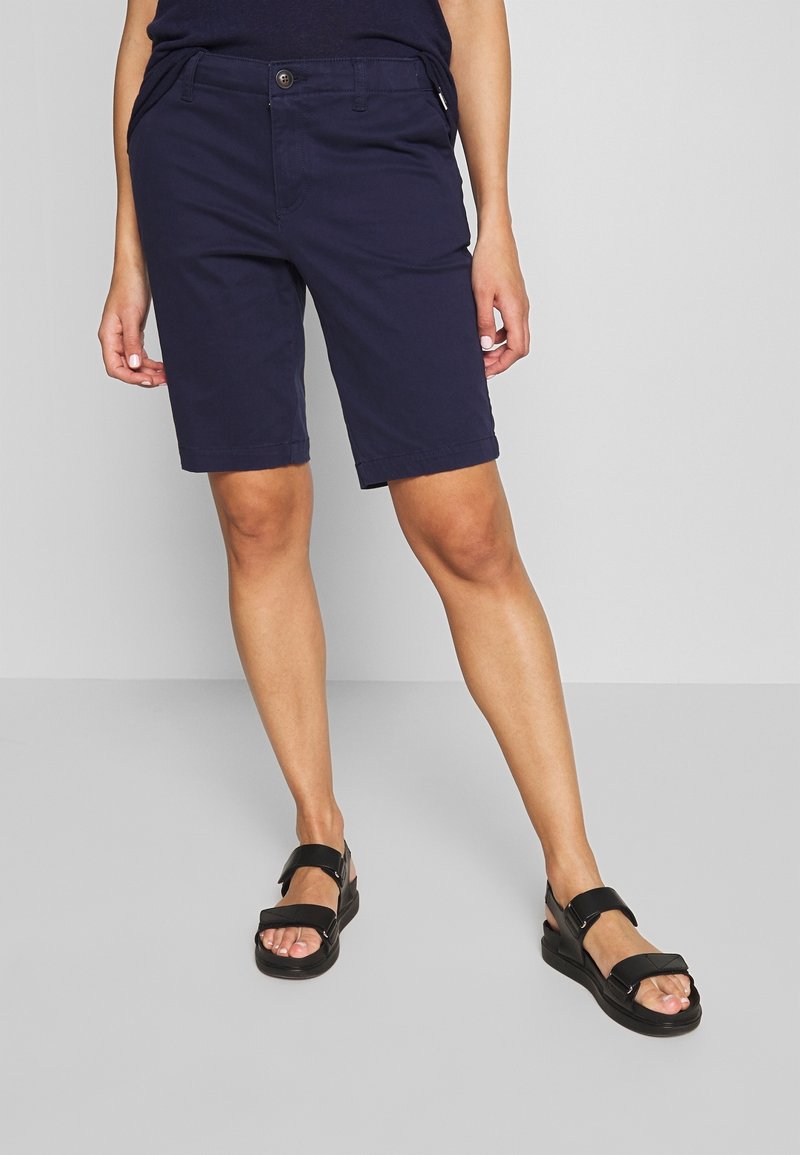 Personne portant un short bleu marine jusqu’aux genoux et des sandales noires ouvertes aux orteils, debout devant un fond uni gris clair.
