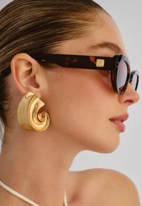 Γυναίκα με μαλλιά που έχουν χτενιστεί προς τα πίσω, φορώντας μεγάλα χρυσά σκουλαρίκια σε σχήμα σπειροειδούς και γυαλιά ηλίου με καφέ ταρταρούγα και τετράγωνο χρυσό λογότυπο στο πόδι.