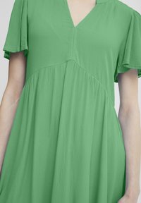 Robe verte avec un décolleté en V, manches courtes et légères, tissu texturé, et silhouette évasée, présentant un détail de couture sur la poitrine.