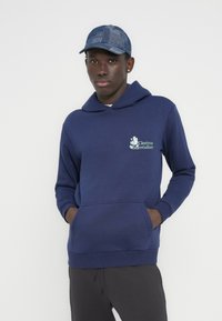 Φούτερ με κουκούλα σε ναυτικό μπλε χρώμα, από απαλό ύφασμα, με μπροστινή τσέπη και λογότυπο που αναγράφει «Cinéma Surréaliste». Φορώντας το με σκουρόχρωμο γκρι παντελόνι και μπλε καπέλο.