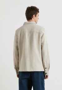 Giovane che guarda verso l'orizzonte, indossando una camicia a maniche lunghe beige testurizzata e pantaloni blu scuro, su uno sfondo chiaro e semplice.