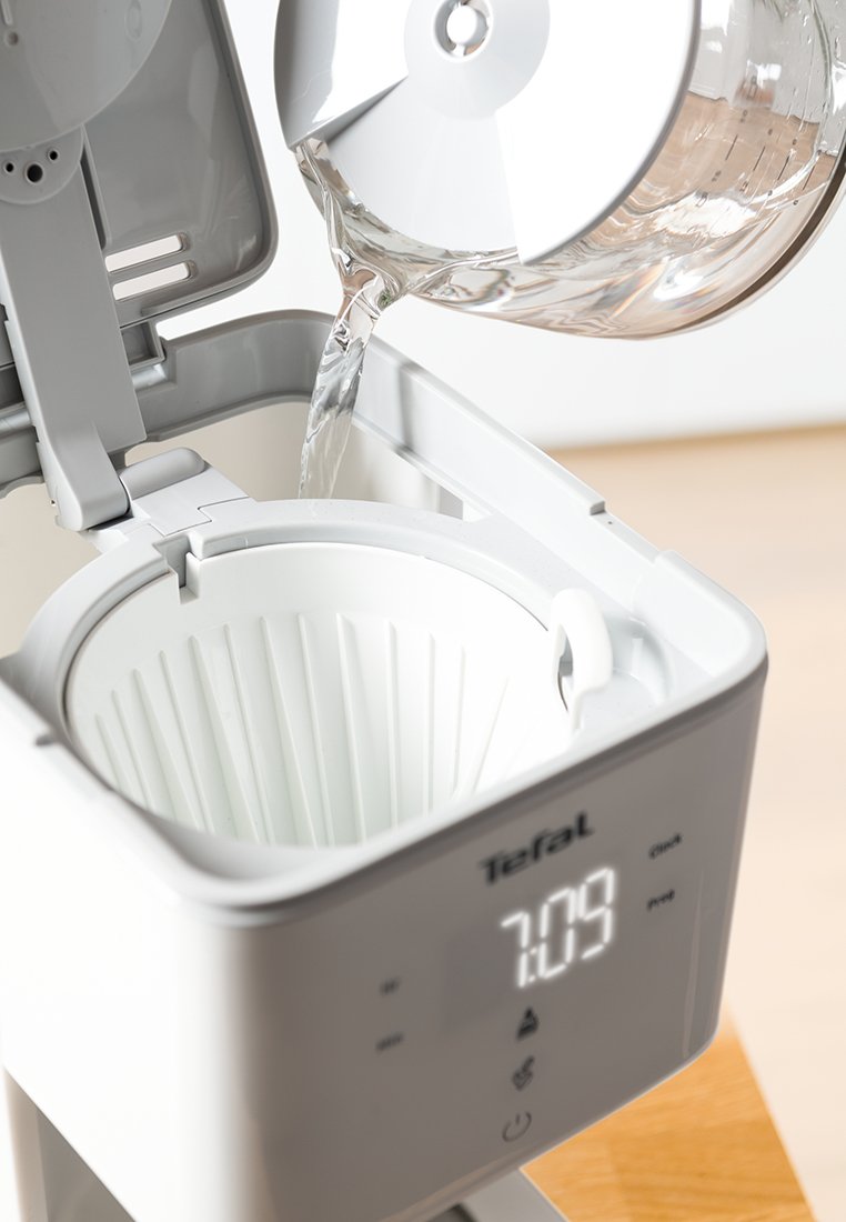 Cafetière électrique avec un boîtier gris, un porte-filtre blanc et une carafe transparente versant de l'eau. L'écran numérique affiche l'heure 7:09.