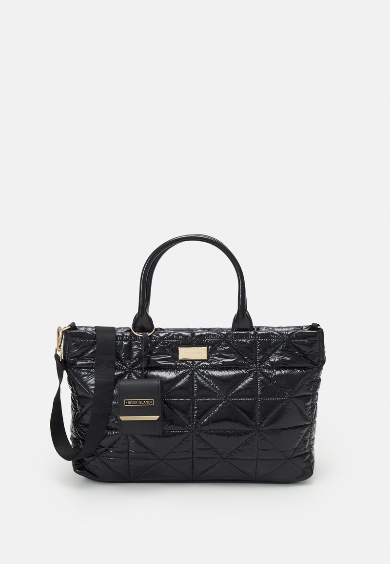 Sac cabas matelassé noir avec double poignée, bandoulière détachable et petite pochette attachée sur fond clair.