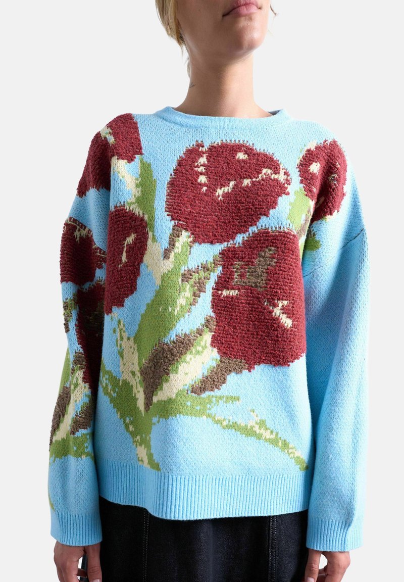 Pull bleu en tricot avec de grandes fleurs rouges et des feuilles vertes porté par une personne devant un fond uni.