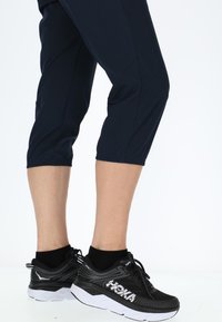 Scarpe sportive nere con dettagli bianchi, indossate con pantaloni navy a vita alta. I pantaloni hanno un orlo elastico; le scarpe presentano una parte superiore testurizzata e una suola imbottita.