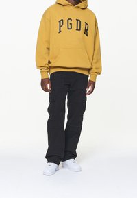Gelber Kapuzenpullover mit der Aufschrift "PGDR" in schwarzer Schrift, kombiniert mit schwarzen Cargohosen mit mehreren Taschen und weißen Turnschuhen.