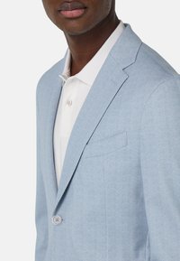 Blazer de espiga azul claro hecho de tela texturizada, con cierre de un botón y solapas muesca sobre una camiseta polo blanca.