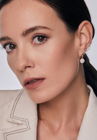 Złote kolczyki w kształcie kół z kwiatowym wzorem oraz przezroczystymi, okrągłymi kamieniami szlachetnymi. Jasne tło podkreśla szczegóły i czyste linie akcesorium.