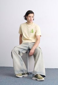 Camiseta color beige con logo verde, combinada con jeans sueltos de color azul claro y zapatillas verde oliva. Modelo sentado en una silla de metal en un entorno neutro.