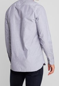 Harmaa, pitkähihainen paita, jossa on hienovarainen tekstuuri, napilliset hihansuut ja kaareva helma. Räätälöity istuvuus takana olevalla yoke-yksityiskohdalla.