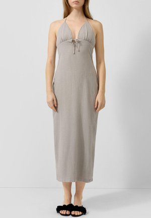 Femme portant une robe midi grise claire sans manches, à col licou, avec un détail noué à l'avant, posant devant un fond gris uni.