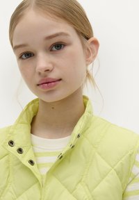 Chaleco acolchado de color amarillo claro con cierre de botones a presión, cuello alto y costuras visibles. El modelo lleva una camiseta de rayas debajo.