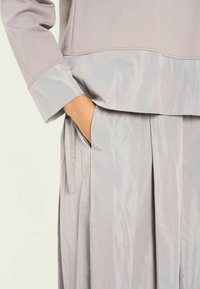 Conjunto en gris claro que presenta una blusa suave y mate, junto con una falda plisada ligeramente brillante. Una mano está introducida en el bolsillo lateral de la falda.