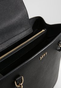 Sac cabas noir en matériau texturé, doté d'une fermeture éclair dorée, de compartiments internes et d'un accent de marque à l'avant.