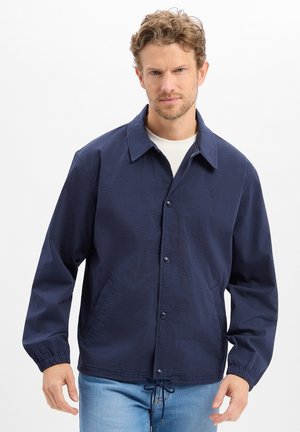 Homme portant une veste boutonnée bleu marine avec des poignets élastiques et une taille à cordon de serrage par-dessus une chemise blanche, sur un fond uni.