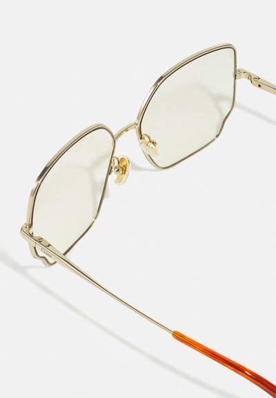 Ochelari de soare din metal aurit cu lentile hexagonale. Lentilele sunt de un verde deschis, cu un accent portocaliu pe brațele ochelarilor. Include suporturi pentru nas transparente.