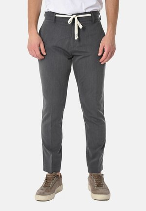 Uomo con pantaloni slim fit grigi con coulisse bianca e sneakers in suede beige in piedi su sfondo bianco