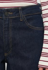 Jean en denim bleu foncé avec une coupe régulière, présentant des coutures contrastantes, des poches avant et un bouton en métal à la taille.