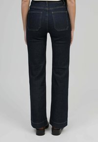 Personne portant un jean droit bleu marine vu de dos, associé à des chaussures à talons marron sur un fond uni.