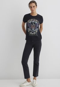 Black cotton t-shirt featuring a colorful butterfly graphic and text; paired with dark jeans and white sneakers. Simple, casual style.