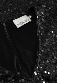 Juodas be rankovių suknelė su V formos iškirpte, padengta blizgiais juodais pailgais sekvinais. Turi diskretišką užtrauktuką ir audinio etiketę su užrašu "Noella S."